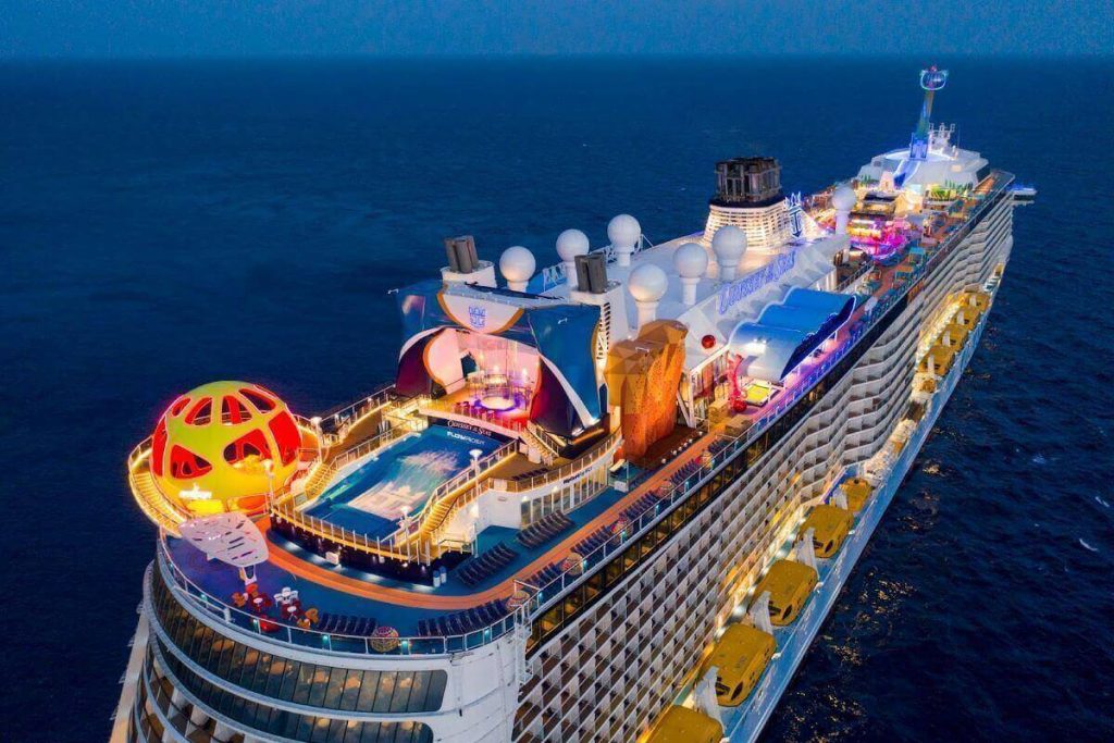 Evening shot of Royal Caribbean's Odyssey of the Seas cruise ship, featuring its deck lit with vibrant lights, including an illuminated sphere structure, outdoor pool, and lounging areas on the calm ocean.