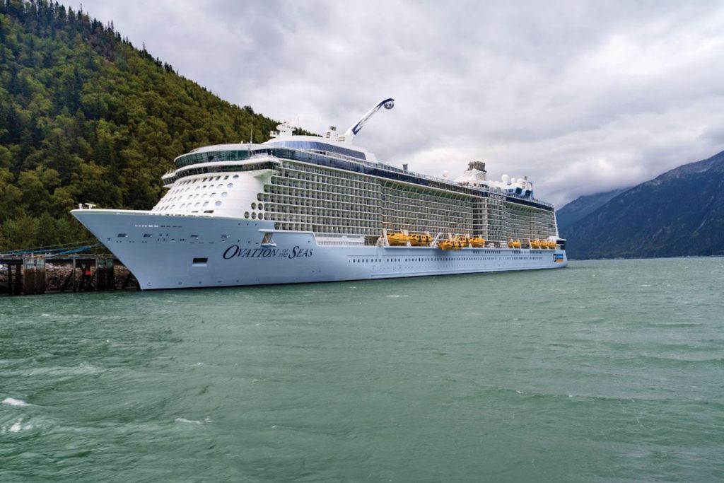 Royal Caribbean's Ovation of the Seas cruise ship moored in a mountainous fjord, its white and blue hull contrasting with the natural greenery and the cloudy sky above the calm glacial waters.