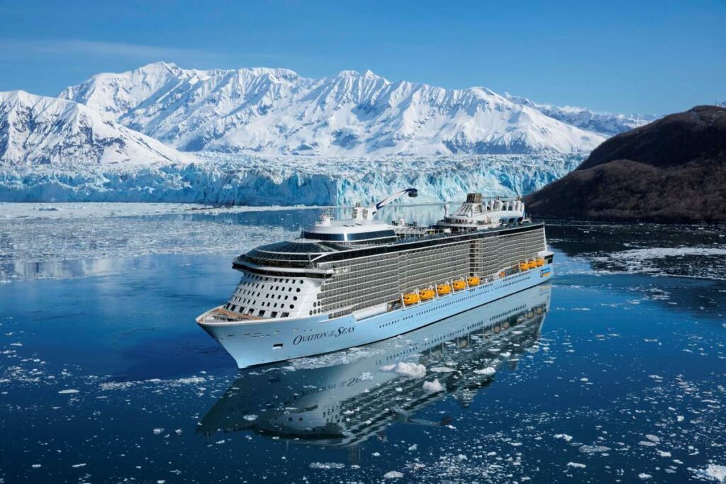 The cruise ship 'Ovation of the Seas' glides through the icy waters of Alaska, with the majestic backdrop of snow-covered mountains and glaciers reflecting the clear blue skies.