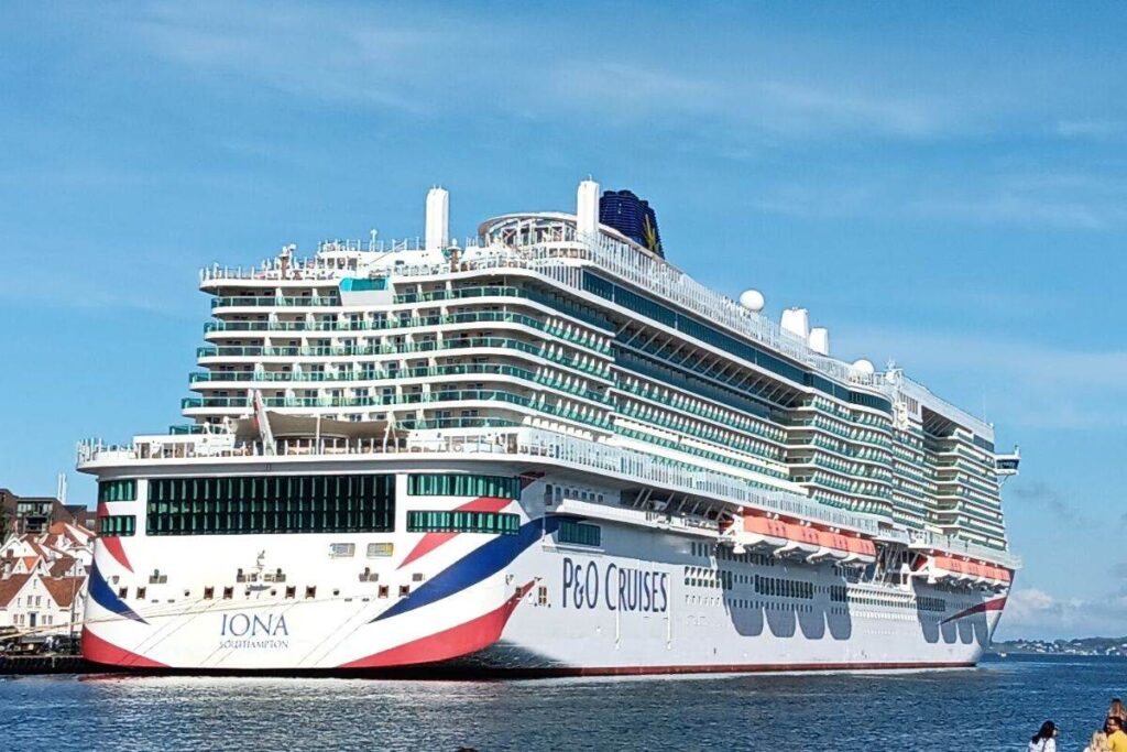 The image shows the P&O Iona cruise ship docked at a port. The ship features a sleek and modern design with multiple decks and large balconies, bearing the P&O Cruises branding and a patriotic red, white, and blue motif at the stern. The backdrop includes clear skies and a coastal town, adding to the scenic view.