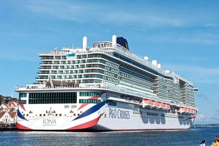The image shows the P&O Iona cruise ship docked at a port. The ship features a sleek and modern design with multiple decks and large balconies, bearing the P&O Cruises branding and a patriotic red, white, and blue motif at the stern. The backdrop includes clear skies and a coastal town, adding to the scenic view.
