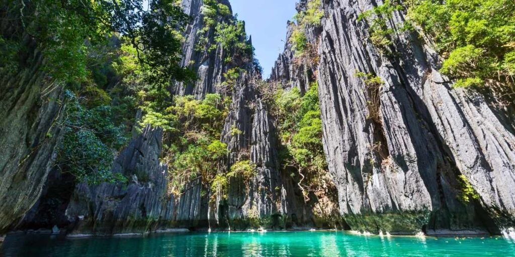 Towering limestone cliffs rise dramatically from crystal-clear turquoise waters in a lagoon in Palawan, Philippines. The rugged rock formations are partially covered with lush greenery, creating a striking contrast against the blue sky.