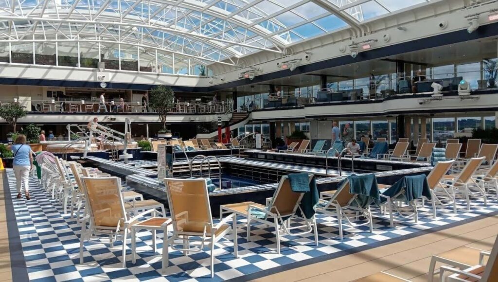 The image showcases the Panorama Pool Club on Deck 9 of Cunard's Queen Anne cruise ship. The pool area features a spacious, glass-covered enclosure allowing for natural light while protecting guests from the elements. Surrounding the pool are numerous loungers with towels neatly draped over them, arranged on a chic checkered floor. The upper deck includes additional seating and dining areas, providing a relaxing atmosphere for guests to enjoy both sun and shade. The design of the space is modern and inviting, perfect for leisurely poolside lounging.