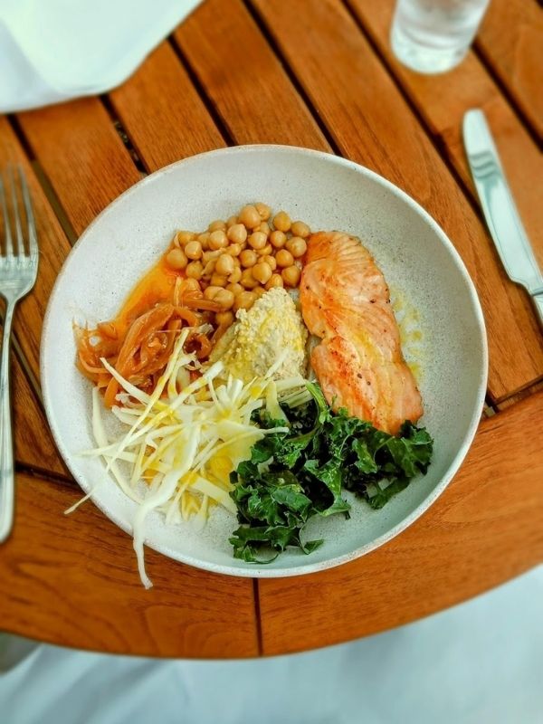 The image shows a healthy meal from the Pavilion Wellness Café on Cunard's Queen Anne cruise ship. The dish is beautifully presented with slices of grilled salmon, chickpeas, sautéed onions, kale, and a serving of creamy hummus, accompanied by shredded vegetables. The meal is plated on a rustic ceramic dish, set on a wooden table, reflecting the café's focus on fresh, nutritious ingredients. This dish embodies a balanced, wellness-focused dining experience.