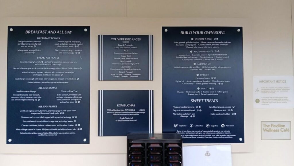 The image shows the menu board at the Pavilion Wellness Café on Cunard's Queen Anne cruise ship. The menu offers a variety of health-conscious options, including breakfast bowls, cold-pressed juices, kombuchas, and build-your-own bowls with a wide selection of bases, ingredients, proteins, and dressings. Sweet treats like vegan chocolate brownies and jam-filled granola cookies are also available, alongside various all-day bowls and plates with fresh ingredients. The menu caters to different dietary preferences, including vegetarian, gluten-free, and plant-based options.
