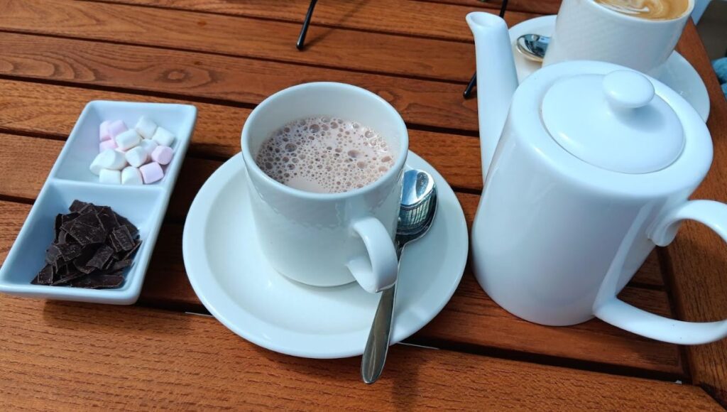 The image shows a delightful hot chocolate setup from the Pavilion Wellness Café on Cunard's Queen Anne cruise ship. A white cup of steaming hot chocolate is served alongside a teapot, with a dish containing small marshmallows and chocolate shavings for adding extra indulgence. The setting is casual yet refined, with the drink placed on a wooden table, creating a cozy and inviting atmosphere for a relaxing moment onboard.