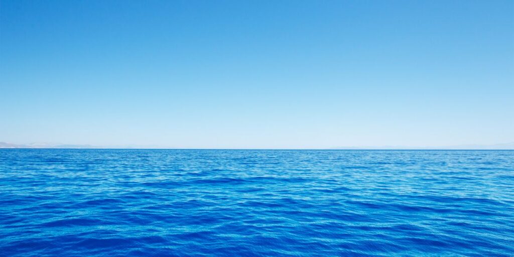 The serene blues of a calm ocean stretch towards a clear sky on the horizon, embodying the peacefulness of a still sea. The simplicity of the scene invites a sense of tranquility and vast open space.