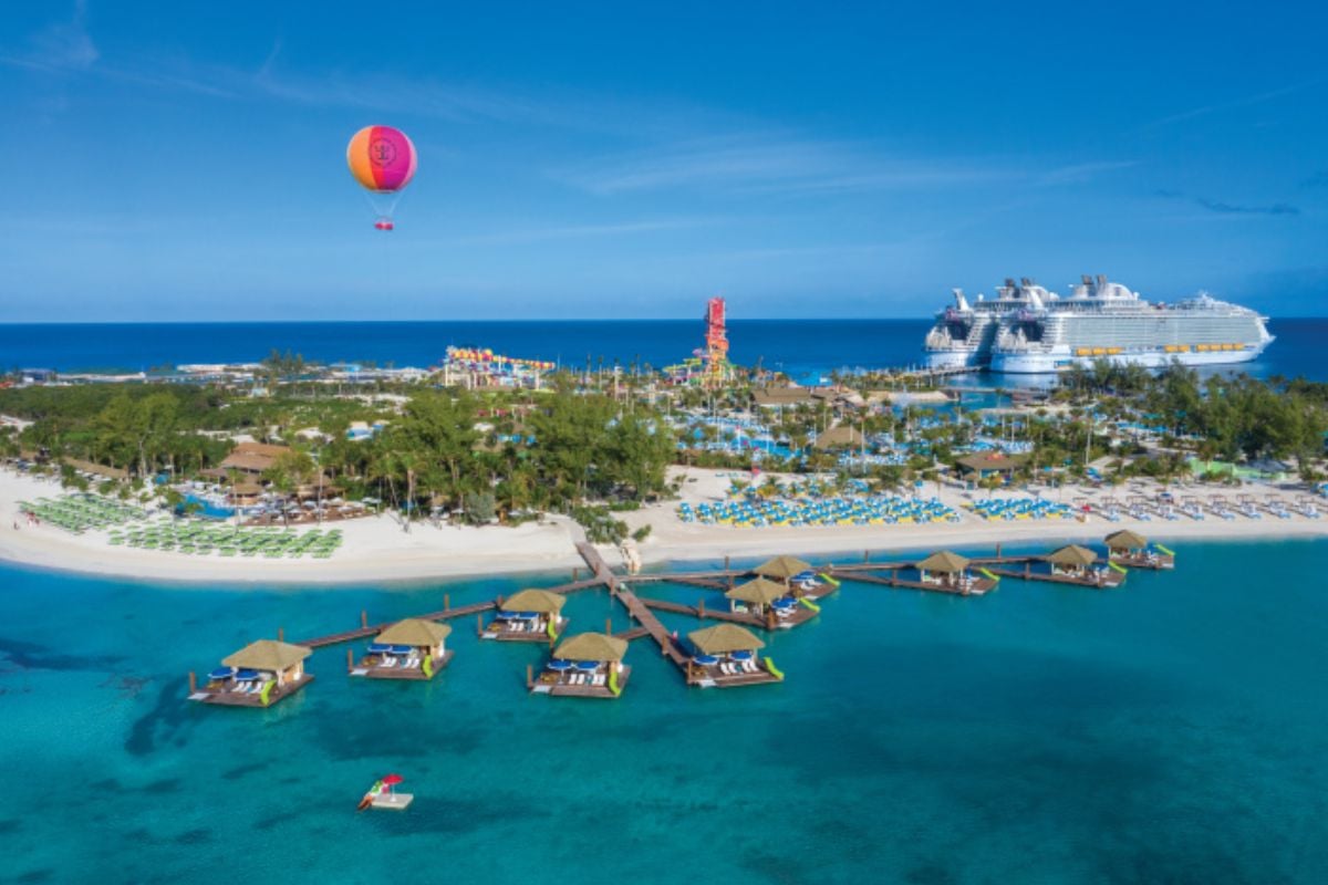 This image captures the vibrant and exciting atmosphere of Perfect Day at CocoCay, a private island resort. The scene showcases colorful beach cabanas over turquoise waters, lush greenery, and a sandy beach filled with rows of lounge chairs. In the background, a water park with towering slides and a large cruise ship docked at the island are visible. A bright orange and pink hot air balloon adds a playful touch as it floats in the clear blue sky, completing the perfect tropical vacation setting.