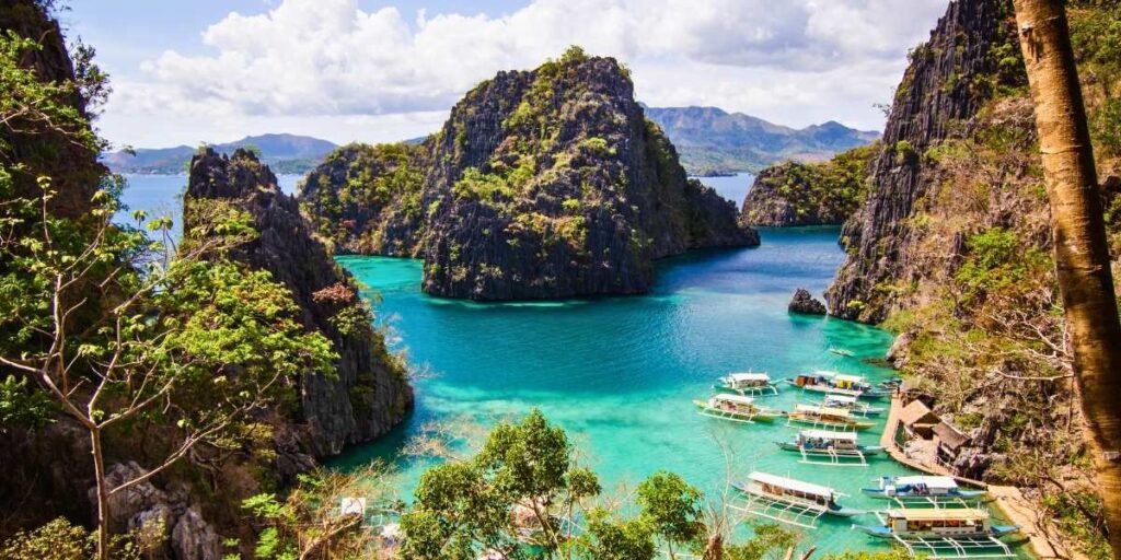 A stunning view of turquoise waters surrounded by dramatic limestone cliffs in Coron, Palawan, Philippines. Several traditional Filipino outrigger boats, known as bangkas, float near a small wooden dock nestled among the lush greenery.