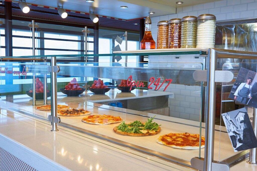 Inside the Pizzeria del Capitano on Carnival Radiance, featuring an array of freshly baked pizzas on display behind a glass counter, with a neon sign stating 'Open 24/7' for round-the-clock dining.