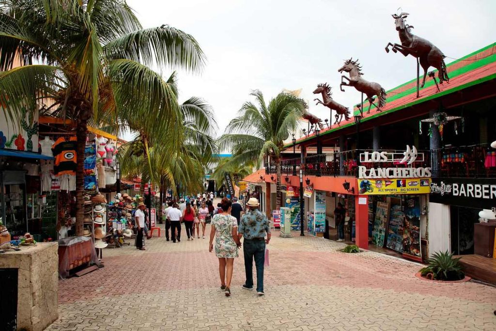 Carnival cruise guests stroll through a vibrant shopping street in Cozumel, lined with palm trees and colorful storefronts, under a cloudy sky.