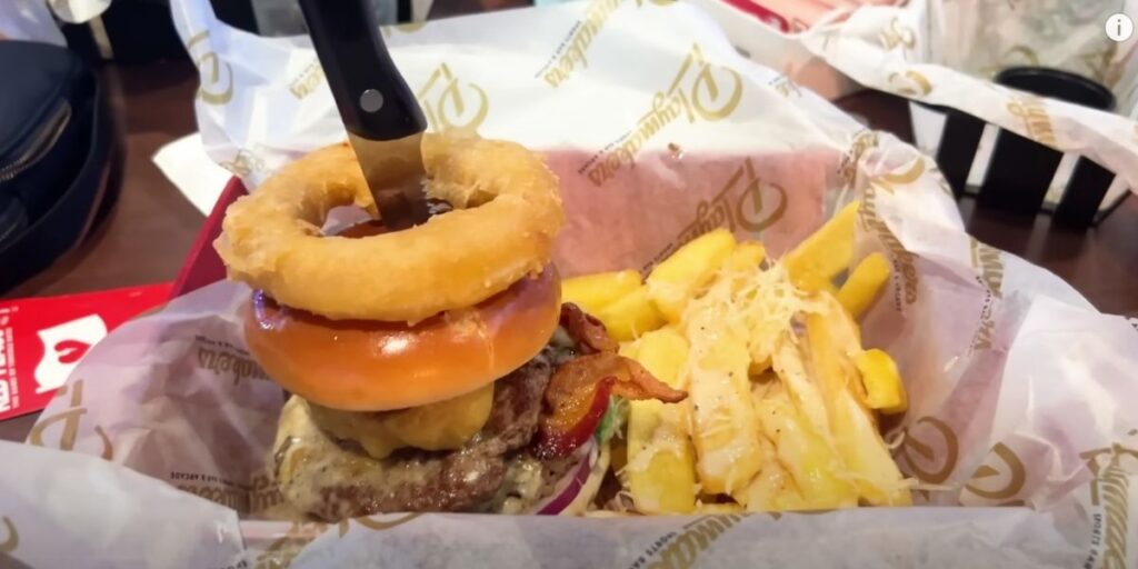 A close-up of a loaded cheeseburger topped with bacon and a crispy onion ring, served with seasoned fries at Playmakers Sports Bar on a cruise ship.