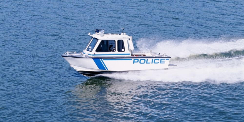 White police patrol boat speeding across open water, creating a trail of mist and waves. The vessel features bold blue stripes and the word “POLICE” clearly marked on the side.