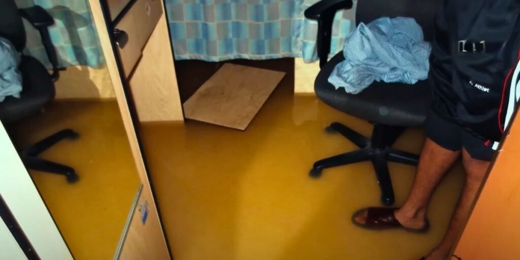 Flooded cruise ship cabin with murky yellowish-brown water covering the floor, partially submerging chairs and furniture; a person's legs are visible standing in the water wearing sandals and shorts, with a striped shirt draped over a chair.