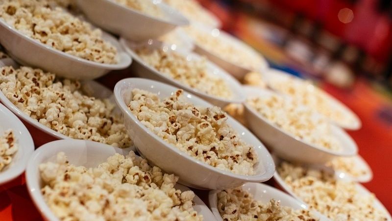 A close-up view of dozens of white bowls filled with freshly popped popcorn, arranged in neat rows on a colorful red table, likely set up for a festive or carnival-style event. The warm lighting enhances the cozy, snack-ready vibe.