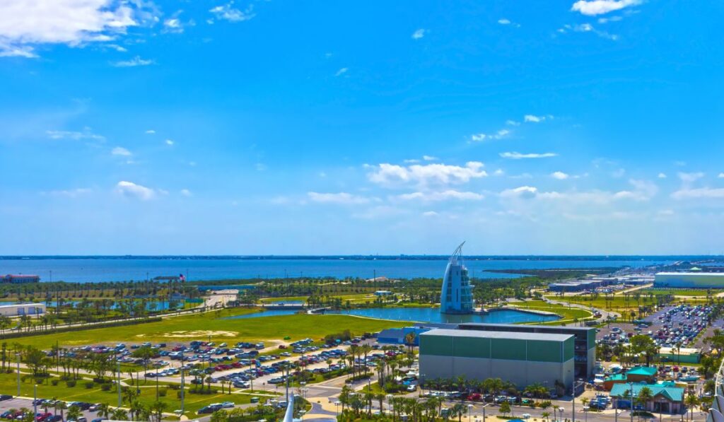 A vibrant view of Port Canaveral, featuring the Exploration Tower with its distinctive architecture against the backdrop of a bright blue sky with scattered clouds. The port's extensive parking area is filled with cars, and the calm waters in the distance reflect the area's role as a gateway for sea travel and exploration.