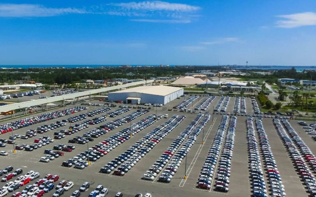 Expansive parking lot at Port Canaveral filled with neatly arranged cars, with industrial buildings in the vicinity and a glimpse of the Florida coastline in the distance under a bright blue sky.