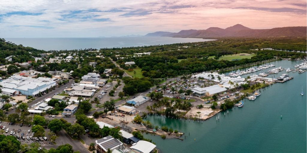 Aerial view of a coastal township adjacent to a marina with moored yachts and surrounded by lush greenery and mountains in the distance. The scene captures the juxtaposition of urban development and natural beauty.