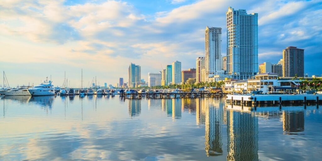 A scenic view of the Port of Manila at sunset, showcasing luxury yachts and sailboats docked in calm waters with the city skyline reflecting on the surface. Tall modern buildings line the coast under a partly cloudy sky, capturing the blend of urban life and seaside charm.