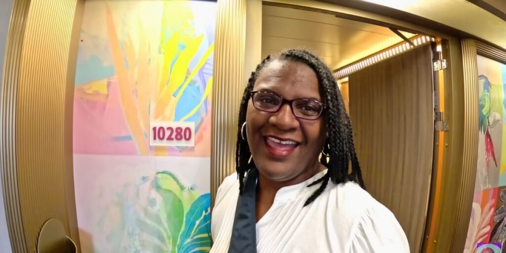 A smiling woman stands in front of her stateroom door on a Carnival cruise ship, with the number "10280" displayed on a colorful, tropical-themed wall panel. She is wearing glasses, a white top, and a black shoulder strap, appearing cheerful and ready for her cruise adventure.