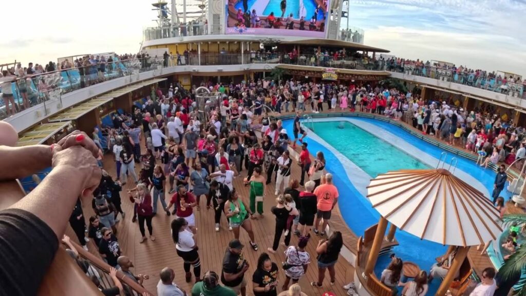 A lively scene on the lido deck of a cruise ship featuring a large crowd of people dancing, mingling, and enjoying a party atmosphere around a central pool. A big screen displays an image of the event, and the deck is surrounded by multiple levels of spectators and seating areas. Bright and festive energy fills the space, with a tiki-style structure in the foreground.