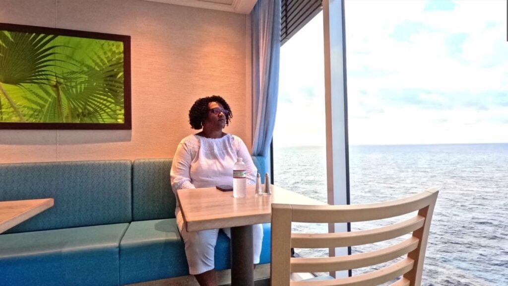 Lynelle from the 'Poverty to Paradise' YouTube channel sitting alone in a cozy dining area by a large window overlooking the ocean. She wears a white top and appears thoughtful, with a water bottle and phone on the table, emphasizing the theme of solo cruising on a budget.