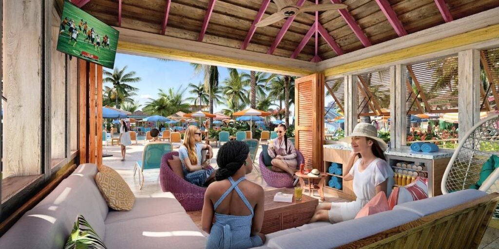 A group of women relaxing inside a colorful premium pool cabana with tropical decor, cushioned seating, and a flat-screen TV showing a soccer game. The cabana overlooks a pool area filled with lounge chairs, umbrellas, and palm trees at Royal Caribbean's Royal Beach Club in Nassau.