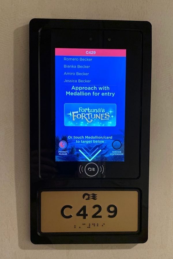 Close-up of a Princess Cruises stateroom door panel for room C429, displaying the names of registered guests and instructions to approach with a Medallion for entry. The digital screen features a game promotion and options to request privacy or service, highlighting the Medallion’s role in keyless cabin access.