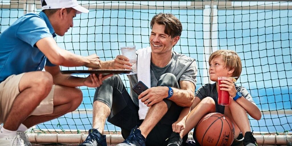 A father and son wearing Princess Cruises Medallion wristbands sit on a basketball court aboard a cruise ship, smiling as a crew member hands them drinks. The relaxed moment highlights personalized service and casual recreation during the voyage.