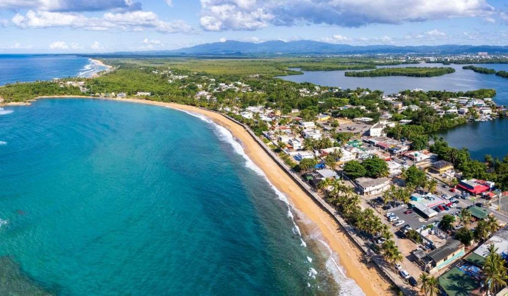 Aerial view of a picturesque curve where the vibrant turquoise Caribbean Sea meets the golden sands of a beach in Puerto Rico, contrasting with the lush greenery and residential areas nearby.