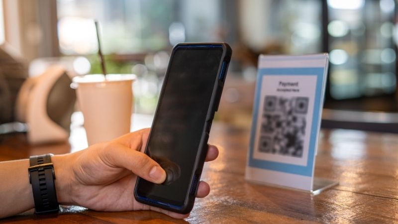 A person holds a smartphone near a tabletop QR code sign labeled "Payment Accepted Here," indicating a contactless digital payment setup in a café setting. A to-go cup with a straw and a payment terminal are visible in the background, reinforcing the casual, tech-savvy vibe.