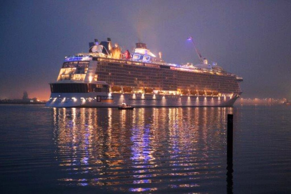The Quantum of the Seas cruise ship, illuminated in the early evening, reflects beautifully on the calm waters at twilight, with lights creating a warm glow and mirrored effect on the surface.