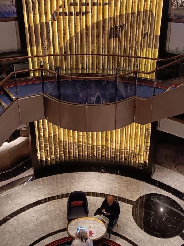 The image depicts a stylish atrium inside Cunard's Queen Anne cruise ship. A golden, illuminated wall with a modern, geometric design serves as the backdrop to a seating area below, where two people are sitting at a round marble table. Above them, a curved staircase with glass railings adds to the elegant design, blending contemporary architecture with luxurious decor. The floors are marble, with a black circular design creating a sense of symmetry and grandeur.
