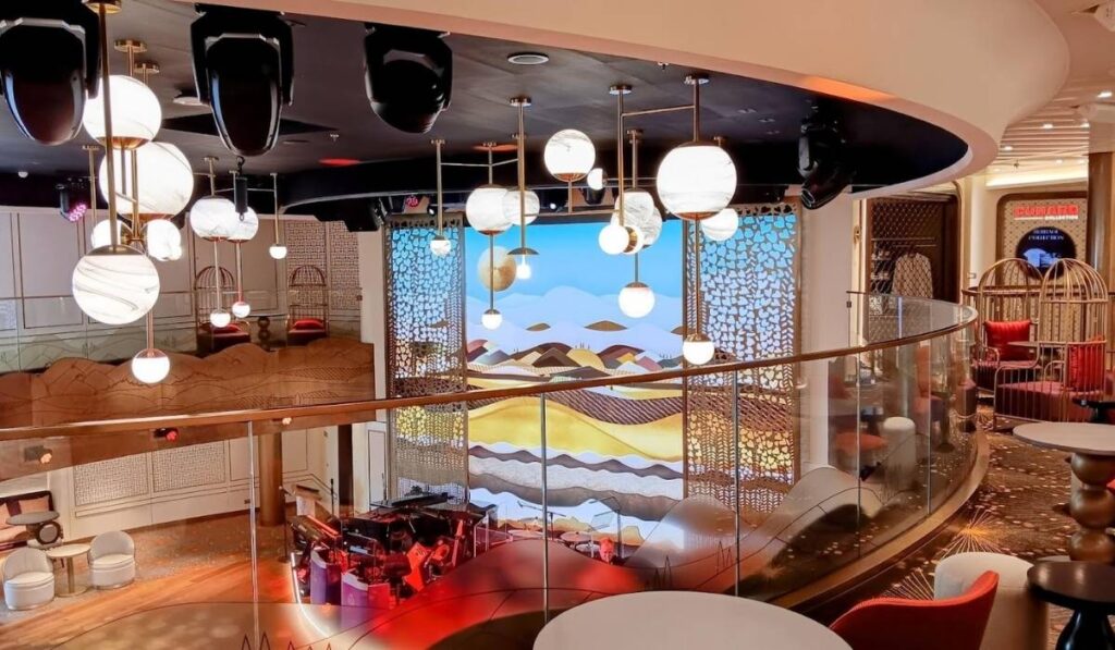 The image shows a modern, artistic lounge area on Cunard's Queen Anne cruise ship. The space is filled with globe-shaped pendant lights hanging from the ceiling, casting a warm glow. In the background, there is a decorative wall with a vibrant, abstract landscape design featuring hills and waves in earthy tones. The seating area is designed with curved lines, cozy red and white chairs, and a glass railing on the upper level. The atmosphere is elegant and inviting, with an emphasis on stylish, contemporary design elements.
