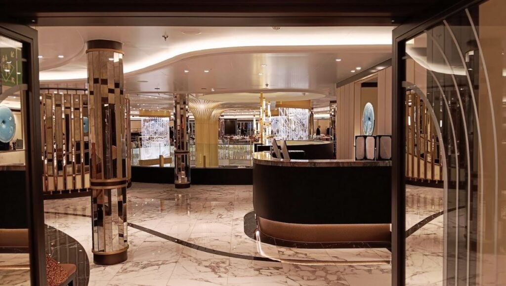 The image shows an opulent interior space on Cunard's Queen Anne cruise ship. The design features glossy marble floors, sleek columns with mirrored accents, and soft, indirect lighting that enhances the luxurious atmosphere. In the distance, decorative metal screens and a striking, column-like structure resembling a fan or tree add to the sophisticated, modern aesthetic. The room feels expansive and elegant, with a clean, refined look, making it a grand and welcoming space for guests on board.
