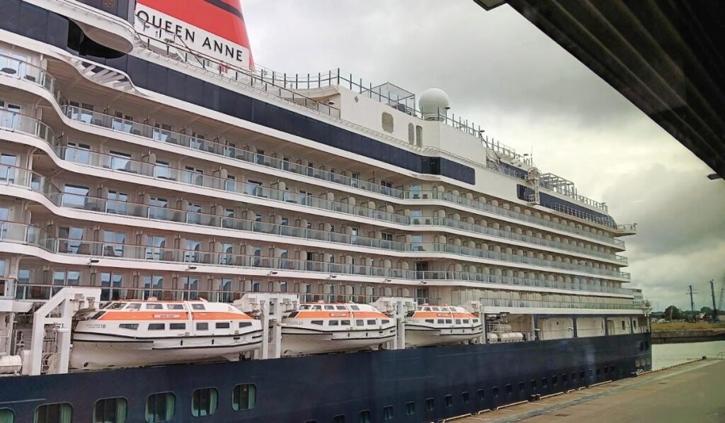 The image shows a side view of Cunard's Queen Anne cruise ship docked at port. The ship's sleek, multi-level design features numerous balcony cabins with glass railings, providing passengers with private outdoor spaces. Several orange and white lifeboats are visible, securely fastened along the side of the vessel. Above the rows of balconies, the ship's name "Queen Anne" is prominently displayed on the iconic red and black funnel. The overcast sky adds a moody atmosphere as the ship rests at the dock, ready for its next journey.