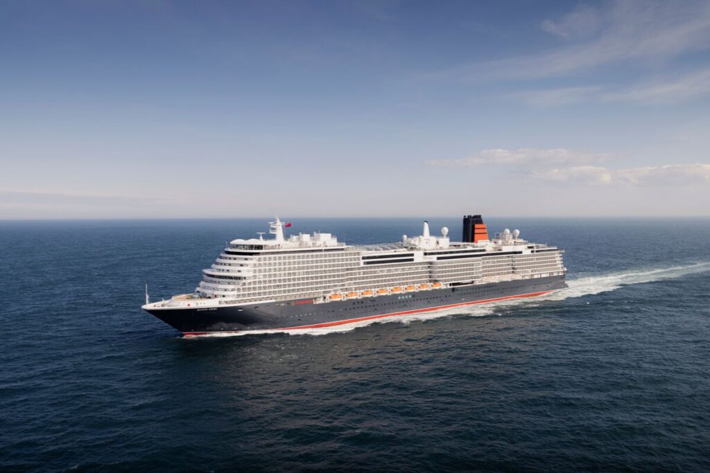 The image shows Cunard's Queen Anne, a large luxury cruise ship, sailing on calm, open waters. The ship has a sleek design, featuring multiple decks and a classic black, red, and white color scheme with orange lifeboats visible along its side. The sky above is clear with a few clouds, suggesting a serene day at sea.