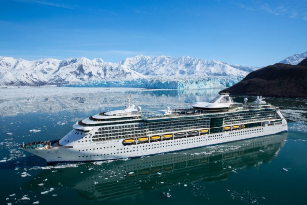 The 'Radiance of the Seas' cruise ship navigates through icy waters with scattered icebergs, set against a backdrop of stunning snow-capped mountains under a clear blue sky.