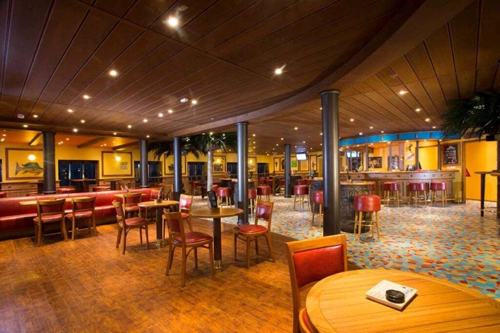 The cozy RedFrog Pub on the Carnival Legend, with its warm wooden floors, red and gold seating, and tropical décor, invites guests to relax and socialize in a comfortable, inviting atmosphere.
