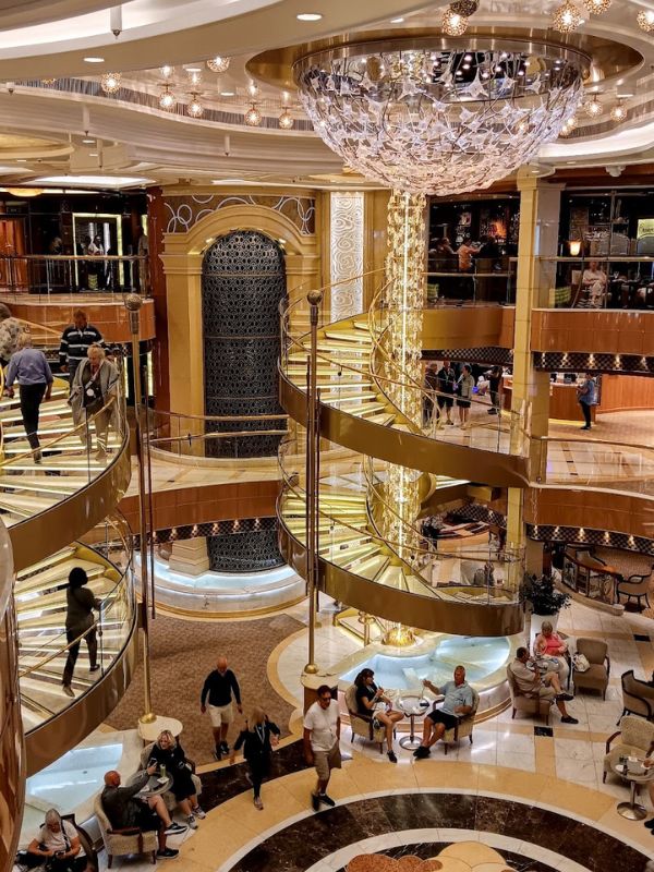 The image shows the luxurious atrium of the Regal Princess cruise ship. The grand interior features a striking central chandelier and spiraling golden staircases with glass railings. Passengers are seen strolling around and relaxing in the seating areas below, enjoying the elegant, well-lit space. The atmosphere is opulent, with intricate designs and a mix of modern and classic décor elements.