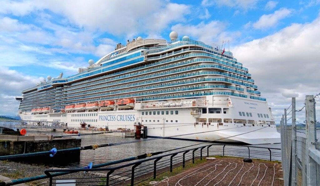 The image shows the Regal Princess, a large, multi-deck cruise ship from **Princess Cruises**, docked at a pier. The ship's sleek white exterior contrasts with the vibrant blue of the many balcony levels, and the lifeboats are visible along its side. The ship is framed against a backdrop of a partly cloudy sky, emphasizing its massive size and luxury features, as it sits calmly by the water.