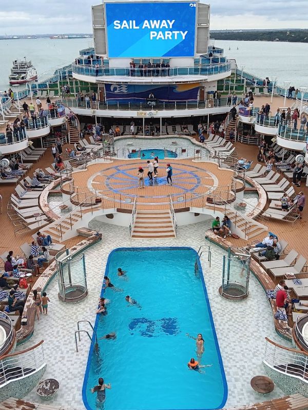 The image depicts the pool deck of the Regal Princess during a "Sail Away Party." Passengers are gathered around the pool, with some swimming and others relaxing on lounge chairs. A large screen overhead displays the event title, adding to the festive atmosphere as the ship prepares to depart from the port. The area is filled with excitement as the cruise begins.