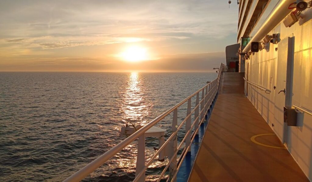 This image shows the Regal Princess cruise ship at sunset, with the sun casting a golden glow over the ocean. The ship's outer deck is visible, with a railing running along the side and reflections of the sunlight on the ship's surface and the water. The peaceful scene highlights the serene atmosphere of cruising at sea during sunset.