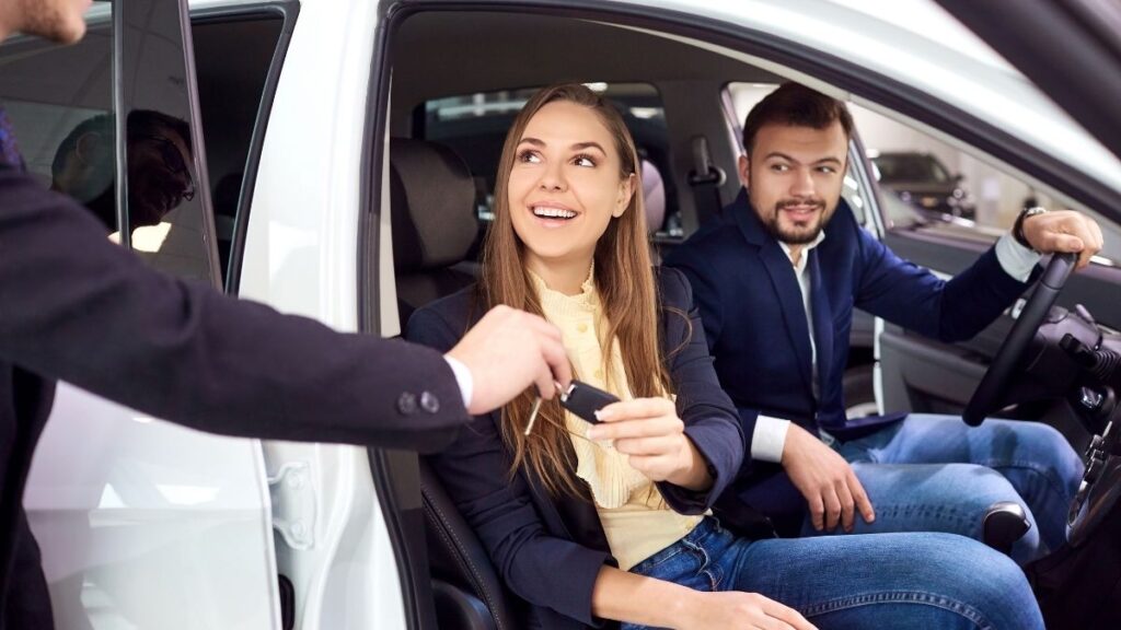 A smiling woman sitting in the passenger seat of a white car reaches out to receive the car keys from a salesman in a suit. A man, dressed in a navy blazer and jeans, sits in the driver’s seat with his hand on the steering wheel, looking on with a pleased expression. The setting appears to be a dealership or rental service, with other vehicles visible in the background.