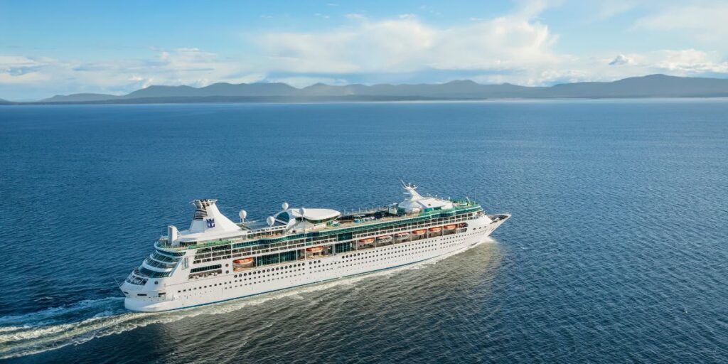 The 'Rhapsody of the Seas' cruise ship from Royal Caribbean International elegantly navigates the calm blue waters with a backdrop of distant mountains under a clear sky. The vessel's white exterior, multiple decks with balconies, and distinctive blue and white funnel are visible, conveying a sense of adventure and luxury travel at sea.