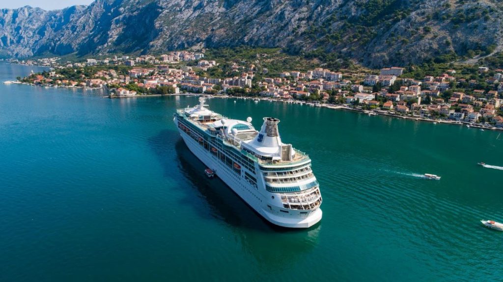 Royal Caribbean's Rhapsody of the Seas cruise ship navigating the emerald waters near a coastal town nestled at the foot of dramatic mountain cliffs, capturing the essence of a Mediterranean getaway.