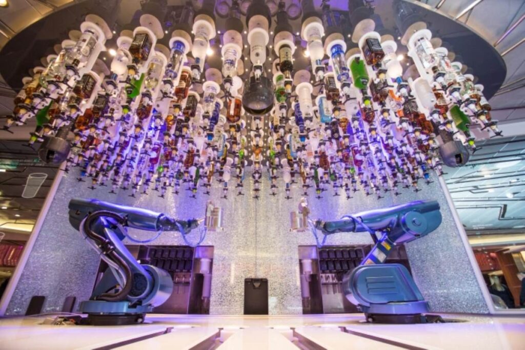 The Robotic Bar on Royal Caribbean's Quantum of the Seas features two robotic arms behind a bar, surrounded by an array of liquor bottles hanging upside down from the ceiling. The robots are programmed to mix and serve drinks to guests, creating a futuristic and innovative bar experience. The setup is illuminated with colorful lights, adding to the high-tech and vibrant ambiance of the venue.