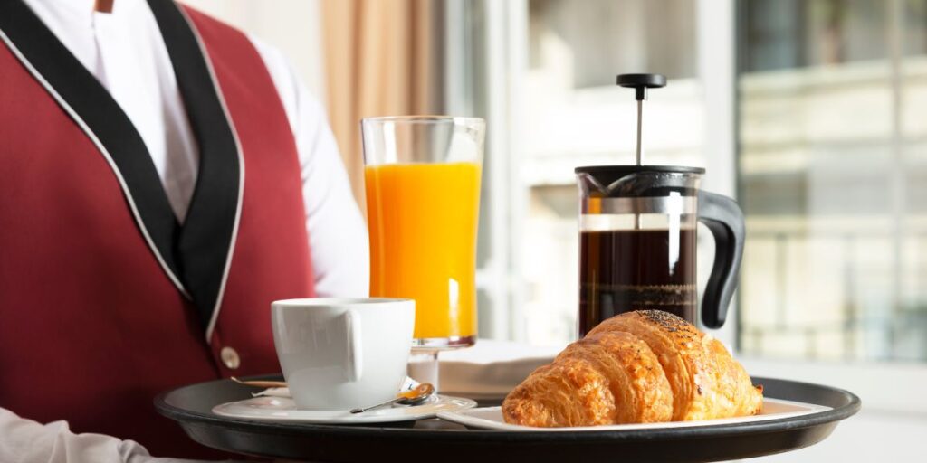 A waiter in a red vest and white shirt serves a breakfast tray featuring a freshly baked croissant, a cup of coffee, a glass of orange juice, and a French press, exemplifying a luxurious and comfortable room service experience with a bright, modern backdrop.