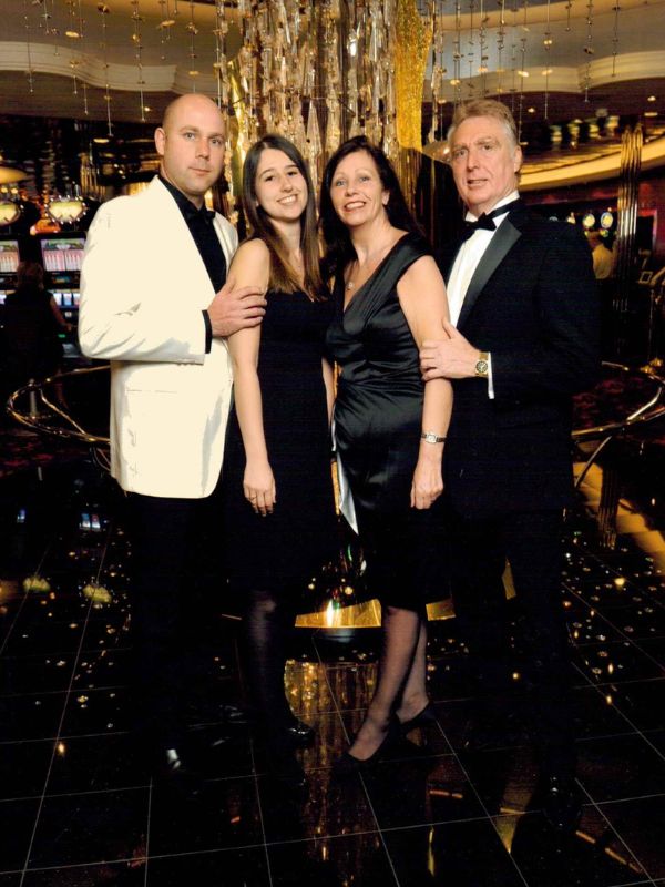 A group of four people, two men and two women, posing for a photo in formal evening wear, with a luxurious chandelier and casino slot machines in the background, indicative of a Royal Caribbean's Oasis of the Seas formal night event.