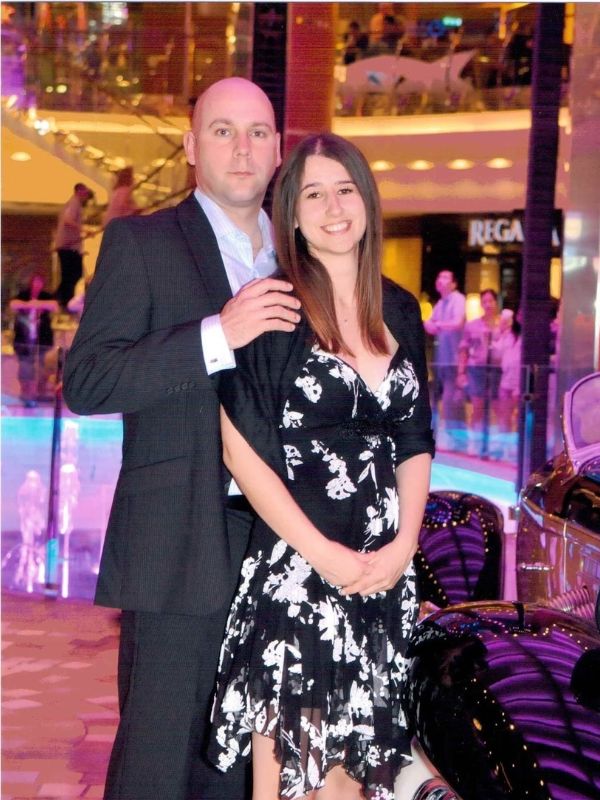A couple embracing each other, dressed in semi-formal attire, with the woman wearing a floral dress, on the Oasis of the Seas cruise ship during a formal night, with a vibrant, illuminated promenade deck in the background.